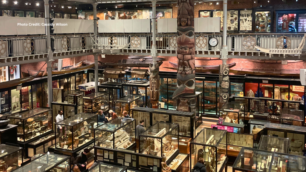 Interior of the Pitt Rivers Museum, featuring numerous glass cases filled with artifacts and a large, iconic totem pole standing tall in the center of the room, reaching to the ceiling.